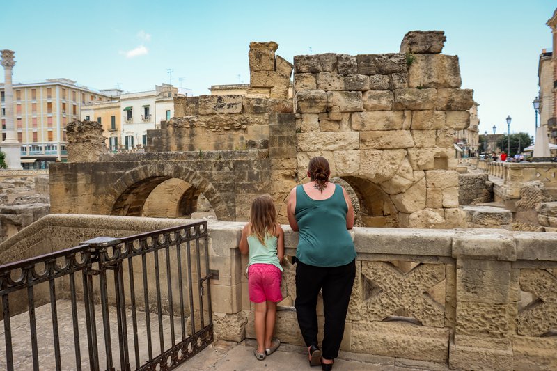 A mom and her daughter lead over a stone wall to look at an ancient structure in Lecce.