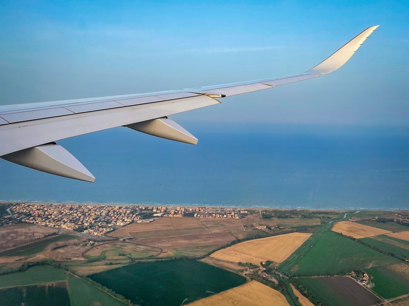 A plane wing extends over an area of Italy, featuring farm land and a city along the sea.