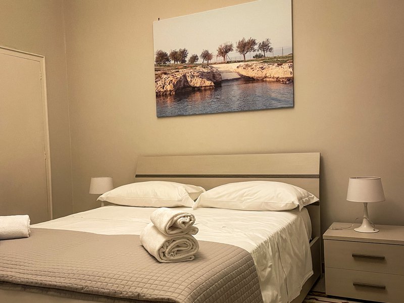A simple bed, with white bedding and towels at the end of the bed, in a simple Vrbo rental in Monopoli, Italy.
